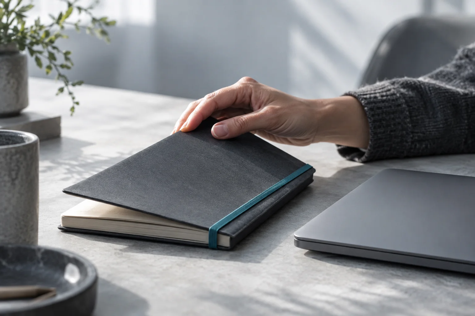 A hand closing a graphite notebook with a turquoise band beside a laptop